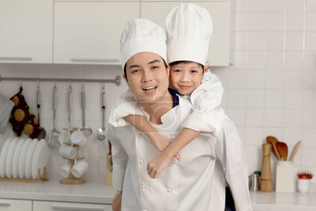 Photo for Happy Asian son kid and father in chef uniform with hat at kitchen, dad piggybacking his kid, parent and boy child playing and having fun together during baking bread, cute family making bakery. - Royalty Free Image