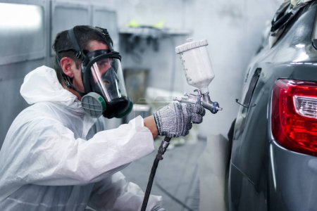 Painting tool in hand of mechanic man painter wearing chemical protective mask while working with auto mechanic painting a car, fixing repairing vehicle at garage automobile repair service shop.