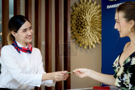 Beautiful smiling Asian female receptionist giving key card to tourist woman customer, female staff at reception counter desk helping female guest for checking in at hotel on holiday vacation.