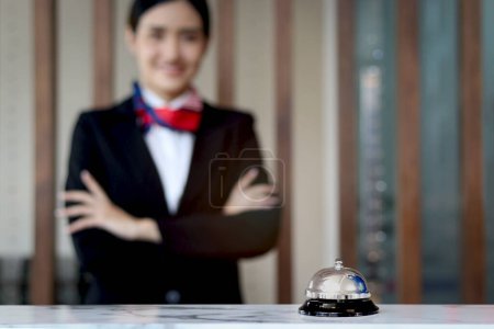 Silver bell on hotel reception service desk with blurred background of smiling beautiful female receptionist in black suit standing with arms crossed, check in hotel service on holiday vacation.