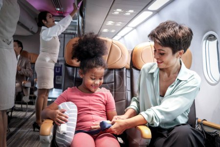 Téléchargez les photos : Le parent prend soin d'un enfant à bord d'un avion, la mère apprend à sa fille à utiliser sa ceinture de sécurité pendant qu'elle est assise à l'intérieur d'un aéronef, une enfant africaine aux cheveux bouclés voyageant en avion, le transport aérien - en image libre de droit