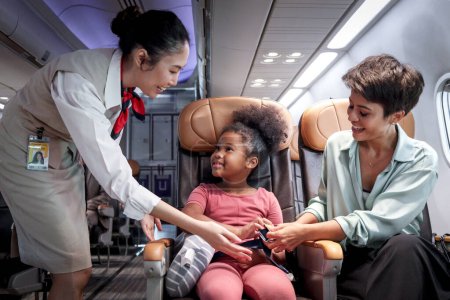 Téléchargez les photos : Hôtesse de l'air amicale prendre soin passager enfant dans l'avion, agent de bord enseigner cheveux bouclés fille africaine à utiliser la ceinture de sécurité pendant assis dans le siège de l'avion, enfant voyageant en avion, transport aérien - en image libre de droit