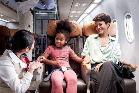 Téléchargez les photos : Hôtesse de l'air amicale prendre soin passager enfant dans l'avion, agent de bord enseigner cheveux bouclés fille africaine à utiliser la ceinture de sécurité pendant assis dans le siège de l'avion, enfant voyageant en avion, transport aérien - en image libre de droit