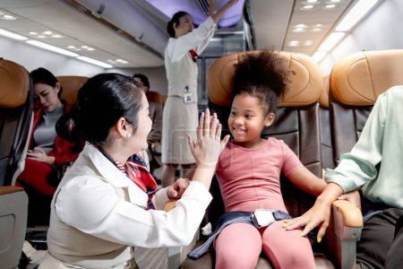 Téléchargez les photos : Hôtesse de l'air amicale prendre soin et aider passager enfant dans l'avion, hôtesse de l'air gaie et cheveux bouclés fille africaine donnant haute cinq sur l'avion, enfant voyageant en avion, transport aérien - en image libre de droit