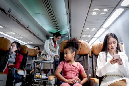 Téléchargez les photos : Heureuse fille africaine enfant et femme passager assis dans un siège confortable à l'intérieur de l'avion pendant hôtesse de l'air femelle poussant chariot sur allée pour servir de la nourriture et des boissons, transport de service aérien - en image libre de droit