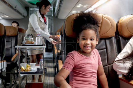 Téléchargez les photos : Happy African fille enfant, enfant passager assis dans un siège confortable à l'intérieur de l'avion pendant femme hôtesse de l'air poussant chariot sur allée pour servir de la nourriture et des boissons, service aérien et le transport - en image libre de droit
