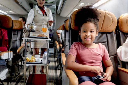 Téléchargez les photos : Happy African fille enfant, enfant passager assis dans un siège confortable à l'intérieur de l'avion pendant femme hôtesse de l'air poussant chariot sur allée pour servir de la nourriture et des boissons, service aérien et le transport - en image libre de droit
