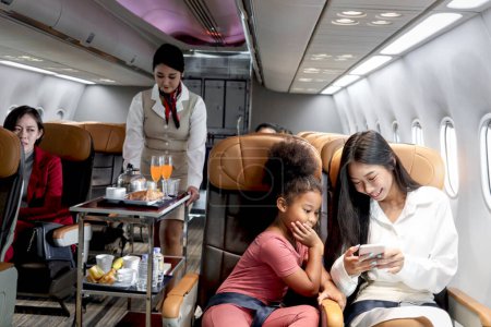 Téléchargez les photos : Heureuse fille africaine enfant et femme passager regardent le téléphone portable pendant assis dans un siège confortable à l'intérieur de l'avion, hôtesse de l'air de cabine féminine poussant chariot sur l'allée pour servir de la nourriture et des boissons - en image libre de droit