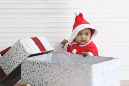 Photo for African baby kid in Santa Claus red costume stands near big gift box present at white room, beautiful little child celebrates Christmas winter holiday. celebration merry Christmas, happy childhood - Royalty Free Image