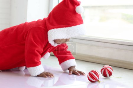 Photo for African baby kid in red Santa Claus costume crawling to grab ball ornament in white living room, beautiful little child girl celebrating Christmas winter holiday. Merry Christmas, happy childhood - Royalty Free Image