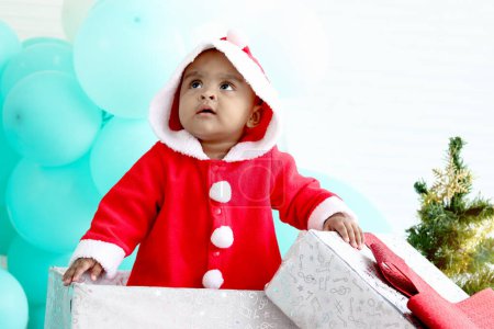 Photo for African baby kid in Santa Claus red costume standing in big gift box present at white room, beautiful little child celebrates Christmas winter holiday. celebration merry Christmas, happy childhood - Royalty Free Image