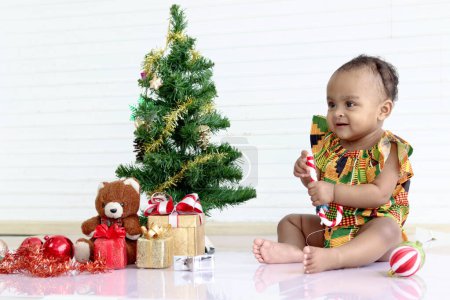 Photo for Cute African baby kid in colorful dress sit near decorative Christmas tree and gift box present in white room, happy cheerful little girl child celebrating happy Christmas winter holiday, happy childhood - Royalty Free Image