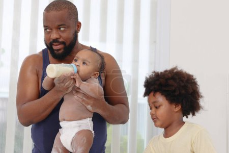 Photo for Happy African family, father feeds toddle baby infant after take a bath and apply talcum powder on body in bathroom. Little daughter child drinking milk from bottle. Dad and brother son look after kid - Royalty Free Image