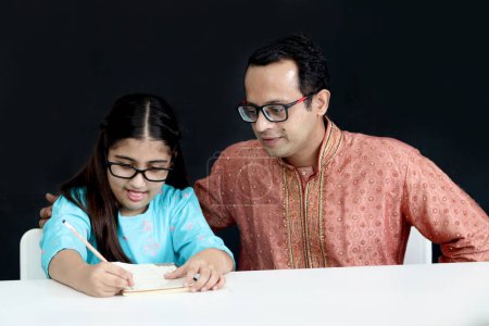 Photo for Cute Indian school student girl in India traditional dress do homework with father. Dad teach daughter kid on black background, parent become teacher tutor involvement in childhood education in family - Royalty Free Image