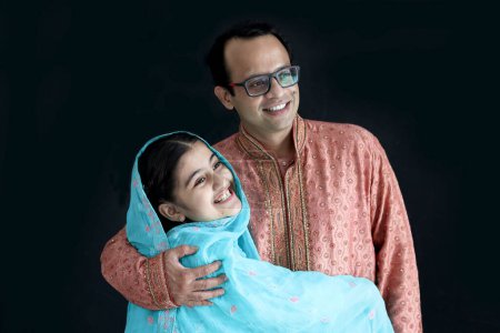 Photo for Cute smiling Indian girl in India traditional blue dress with her father standing on black background, daughter kid hugging her dad with love. Portrait of happy Indian family, father with child. - Royalty Free Image