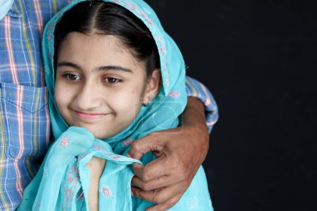 Photo for Portrait of happy smiling Indian Muslim girl child in India traditional dress wearing hijab scarf hugging her father on black background. Daughter kid girl with beautiful eyes in blue costume. - Royalty Free Image
