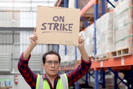 Foto de Enojado trabajador senior asiático infeliz usando chaleco de seguridad, sosteniendo y levantando un cartel en la pancarta de huelga en el almacén logístico de carga. Trabajadores en huelga protestando en el lugar de trabajo. - Imagen libre de derechos