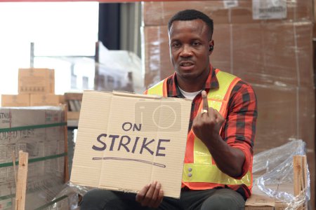 Foto de Enojado trabajador africano infeliz usando chaleco de seguridad y dando el dedo medio con cartel de la pancarta de huelga en el almacén logístico de carga. Trabajadores en huelga protestando en el lugar de trabajo. - Imagen libre de derechos