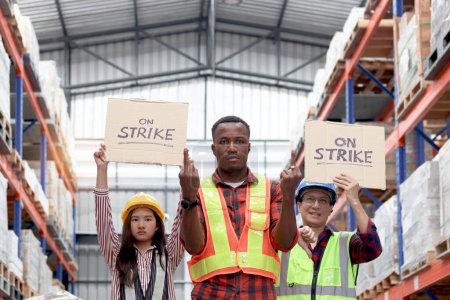 Foto de Enojado trabajador africano infeliz con colegas, personal de alto nivel asiático y mujeres usan chaleco de seguridad y casco, mantenga un cartel en la bandera de huelga en el almacén logístico de carga. Trabajadores en huelga protestando en el lugar de trabajo - Imagen libre de derechos
