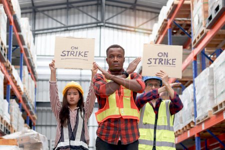 Foto de Enojado trabajador africano infeliz con colegas, personal de alto nivel asiático y mujeres usan chaleco de seguridad y casco, mantenga un cartel en la bandera de huelga en el almacén logístico de carga. Trabajadores en huelga protestando en el lugar de trabajo - Imagen libre de derechos