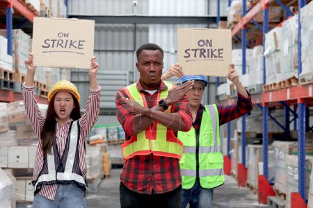 Photo for Angry unhappy African worker with colleagues, Asian senior and woman staffs wear safety vest and helmet, hold sign on strike banner at cargo logistic warehouse. Striking worker protesting at workplace - Royalty Free Image