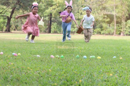 Colorful Easter eggs on green grass backyard with blurred background children with bunny ears holding basket, running to pick up Easter eggs. Kids celebrating Easter spring holiday at outdoor garden.