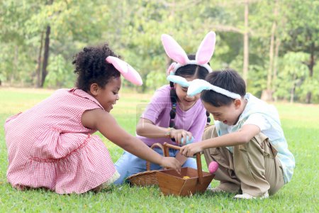 Three little children with bunny ears have fun in park. Asian girl and boy with curly hair African child friend hunting Easter eggs in green garden. Kids celebrating Easter spring holiday at outdoor.