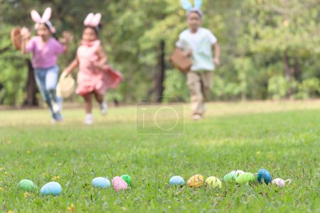 Colorful Easter eggs on green grass backyard with blurred background children with bunny ears holding basket, running to pick up Easter eggs. Kids celebrating Easter spring holiday at outdoor garden.
