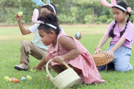 Three little children with bunny ears have fun in park. Curly hair African girl with Asian girl and boy friend hunting Easter eggs in green garden. Kids celebrating Easter spring holiday at outdoor.