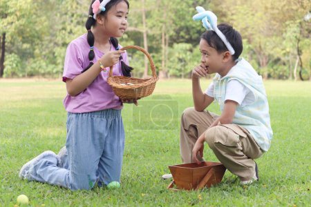 Two little children friend with bunny ears have fun in park. Asian boy and girl hunting and picking up Easter eggs in green garden. Kids celebrating Easter spring holiday at outdoor.