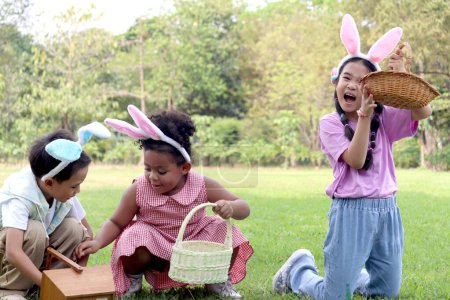 Children with bunny ears have fun in park. Asian girl showing Easter egg basket while boy with curly hair African child friend hunting Easter eggs in green garden. Kids celebrating Easter at outdoor.