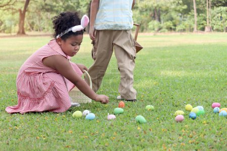 Little children with bunny ears have fun in park. Curly hair African girl with  friend hunting and picking up Easter eggs in green garden. Kids celebrating Easter spring holiday at outdoor.