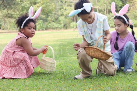 Three little children with bunny ears have fun in park. Curly hair African girl with Asian girl and boy friend hunting Easter eggs in green garden. Kids celebrating Easter spring holiday at outdoor.