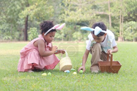 Two little children with bunny ears have fun in park. Curly hair African girl with Asian boy friend hunting Easter eggs in green garden. Kids celebrating Easter spring holiday at outdoor.