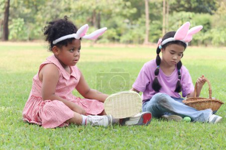 Two little children with bunny ears have fun in park. Curly hair African girl with Asian girl friend hunting Easter eggs in green garden. Kids celebrating Easter spring holiday at outdoor.