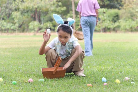 Cute Asian boy with bunny ears finding and hunting Easter eggs in green garden with friends, little children have fun in park. Kids celebrating Easter spring holiday at outdoor.