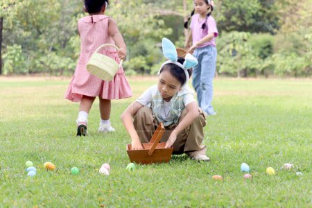 Cute Asian boy with bunny ears finding and hunting Easter eggs in green garden with friends, little children have fun in park. Kids celebrating Easter spring holiday at outdoor.
