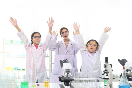 Glückliche zwei junge asiatische Studentinnen im Laborkittel mit Wissenschaftlerinnen, die die Hände heben, um den Erfolg des naturwissenschaftlichen Experiments im Schullabor zu feiern. Kinder lernen naturwissenschaftliche Ausbildung.
