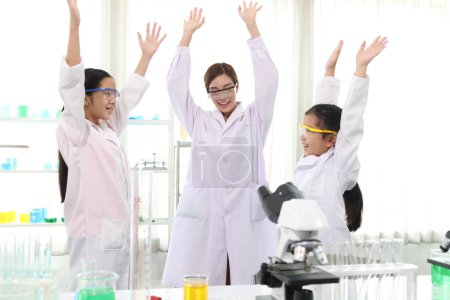 Glückliche zwei junge asiatische Studentinnen im Laborkittel mit Wissenschaftlerinnen, die die Hände heben, um den Erfolg des naturwissenschaftlichen Experiments im Schullabor zu feiern. Kinder lernen naturwissenschaftliche Ausbildung.