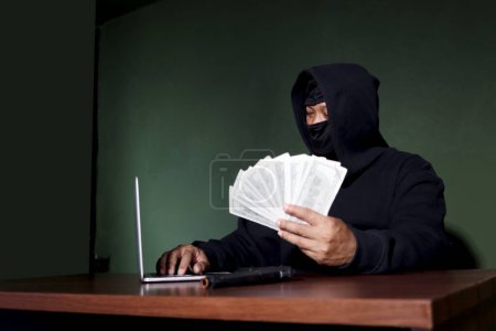 Criminal man in black hood holding money banknotes (mockup) on hand, looking at laptop computer at desk, mask thief robber committing committed cybercrimes online