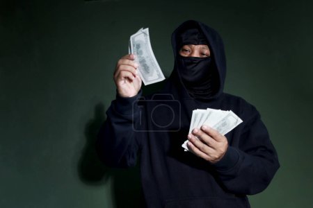 Criminal man in black hood holding banknotes (mockup) and counting money in hand, mask thief robber committing committed cybercrimes online concept.