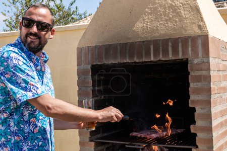 Téléchargez les photos : Young hipster man with a beard, sunglasses and hawaiian shirt drinking a beer while preparing a barbecue to eat at the holiday chalet on summer vacation - en image libre de droit