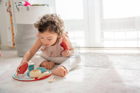 Little 2-year-old girl plays sitting on the floor of her room with a wooden toy that simulates tea and coffee. montessori material concept of learning, development and skills in the childhood stage.