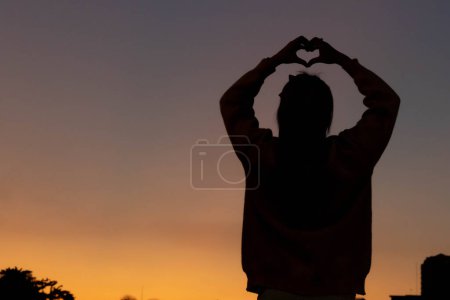 A silhouette of a young woman raising her hands above her head to represent a heart symbol signifying friendship, love and kindness. Heart symbol concept with the meaning of love and friendship.
