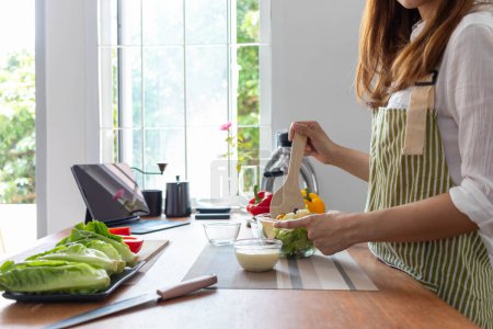 Foto de Mujer joven está haciendo ensalada de verduras que ha preparado en la mesa en su cocina casera para obtener ensalada que es limpia y segura porque los ingredientes se seleccionan cuidadosamente. ideas de preparación de alimentos saludables - Imagen libre de derechos
