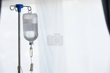 An iron stand to hang saline bottle high to deliver saline via catheter to an intravenous patient lying on patient bed. Medical concept in which doctor gives saline solution to patient through vein