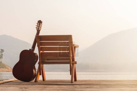 Die hölzerne Gitarre wurde morgens neben einem Stuhl auf einem hölzernen Balkon über einem Stausee mit herrlichem Blick auf die Natur und strahlendem Sonnenschein aufgestellt, um sich auf die bevorstehende Party vorzubereiten..