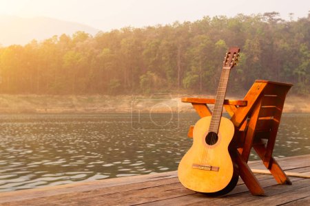 Die hölzerne Gitarre wurde morgens neben einem Stuhl auf einem hölzernen Balkon über einem Stausee mit herrlichem Blick auf die Natur und strahlendem Sonnenschein aufgestellt, um sich auf die bevorstehende Party vorzubereiten..