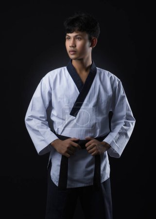 Male black belt taekwondo instructor in white uniform standing pose holding black belt on black background