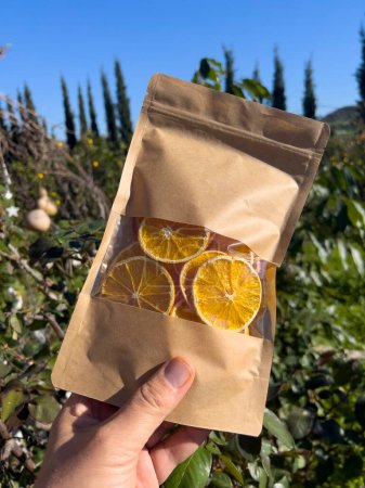 Foto de Natural dried orange chips in brown recycled paper food bag with valve and seal hold by human hand under sun light on green nature background with copy space. Blank no name material, close-up photo. - Imagen libre de derechos